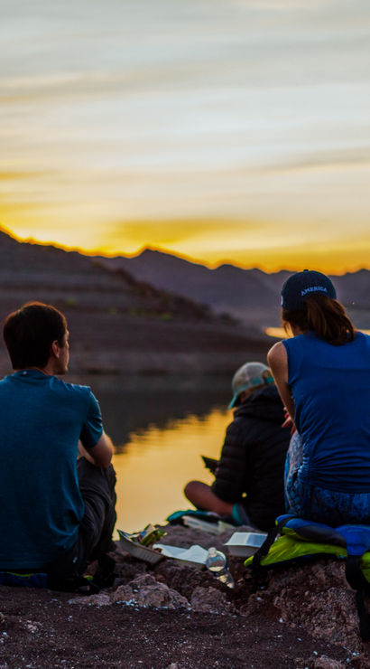 Group of people sitting on a rocky lakeshore watching a golden sunset over mountain ridges, calm water reflecting the colorful sky