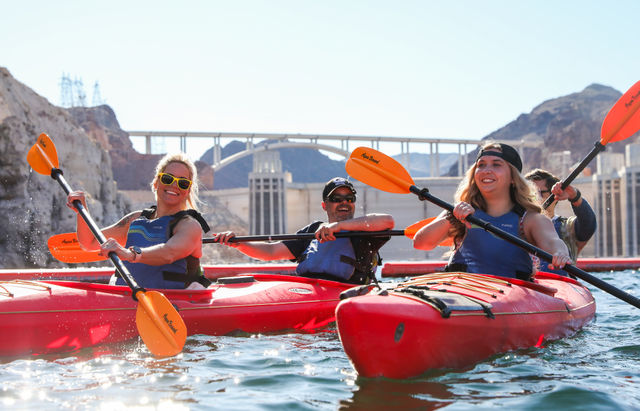 Three smiling kayakers in red kayaks paddling near Hoover Dam and the arched bridge on a sunny day, splashing water and wearing life vests and sunglasses.