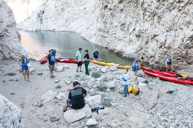 Group prepping colorful red and yellow kayaks on a rocky canyon riverbank beneath steep pale cliffs with calm green water — outdoor kayaking adventure.