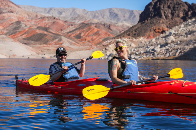 Two people in life jackets smiling and paddling a bright red tandem kayak with yellow paddles on a blue desert lake framed by red‑rock cliffs under clear skies.