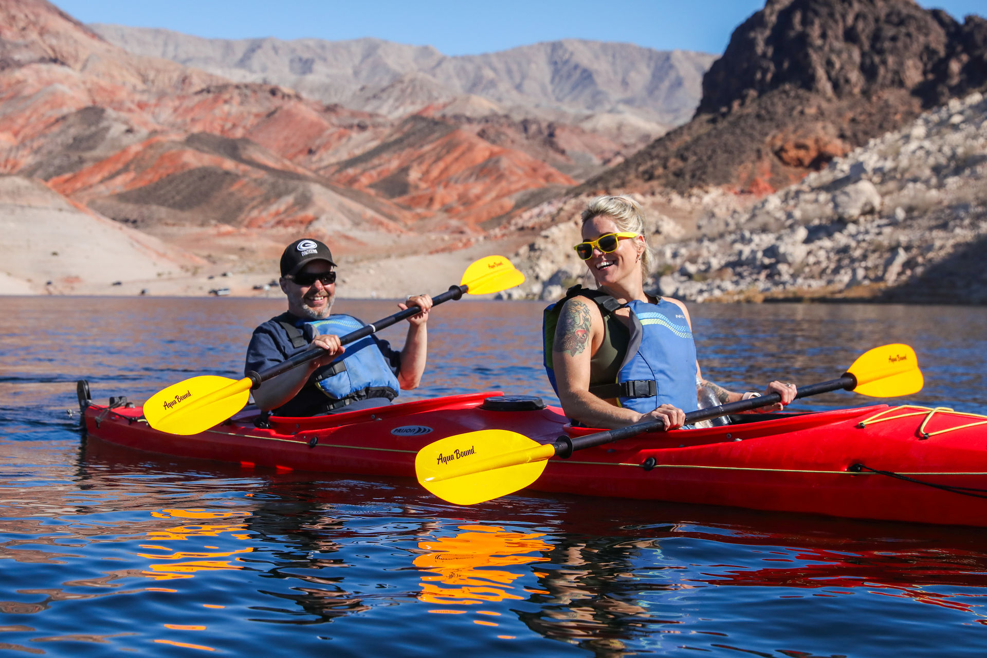 Two people in life jackets smiling and paddling a bright red tandem kayak with yellow paddles on a blue desert lake framed by red‑rock cliffs under clear skies.