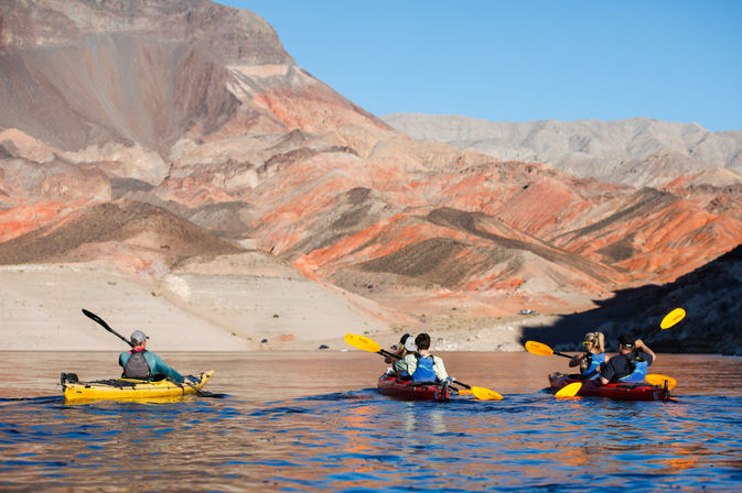 Four kayakers paddling on a calm blue desert lake in bright yellow and red kayaks, set against layered red-orange canyon slopes under a clear blue sky.