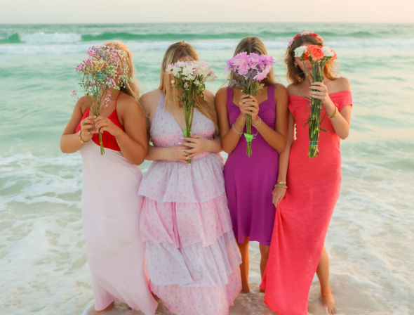 Four people in bright summer dresses stand ankle-deep on a pastel beach at sunset, each hiding behind a colorful bouquet with turquoise waves rolling in the background.