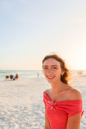 Smiling woman wrapped in a pink towel with a plumeria flower, standing on a sunlit sandy beach at sunset with the ocean and beachgoers in the background.
