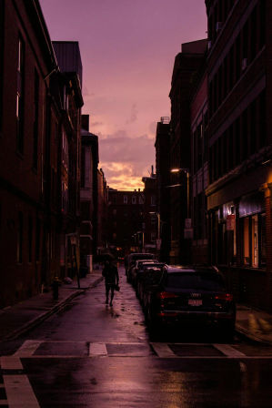 Moody purple-orange sunset over a wet downtown city street — silhouette of a person with an umbrella walking past parked cars between brick buildings, streetlights reflecting on the pavement.