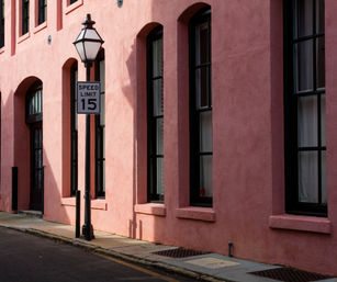Charming pink stucco building facade with arched black‑trim windows, vintage streetlamp and a Speed Limit 15 sign along a narrow city sidewalk.
