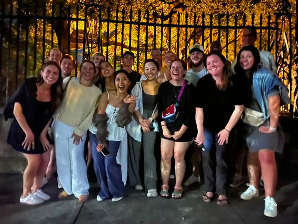 Group photo of about 15 friends smiling on a city sidewalk at night, posing in front of a decorative iron fence backlit by warm tree lights.
