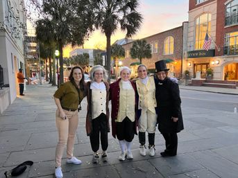 Five people smiling on a palm-lined historic downtown street at sunset — four in colonial-era costumes and one in casual clothes, with brick buildings, lampposts and an inn flying an American flag in the background.