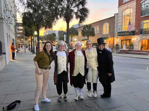 Five people smiling on a palm-lined historic downtown street at sunset — four in colonial-era costumes and one in casual clothes, with brick buildings, lampposts and an inn flying an American flag in the background.