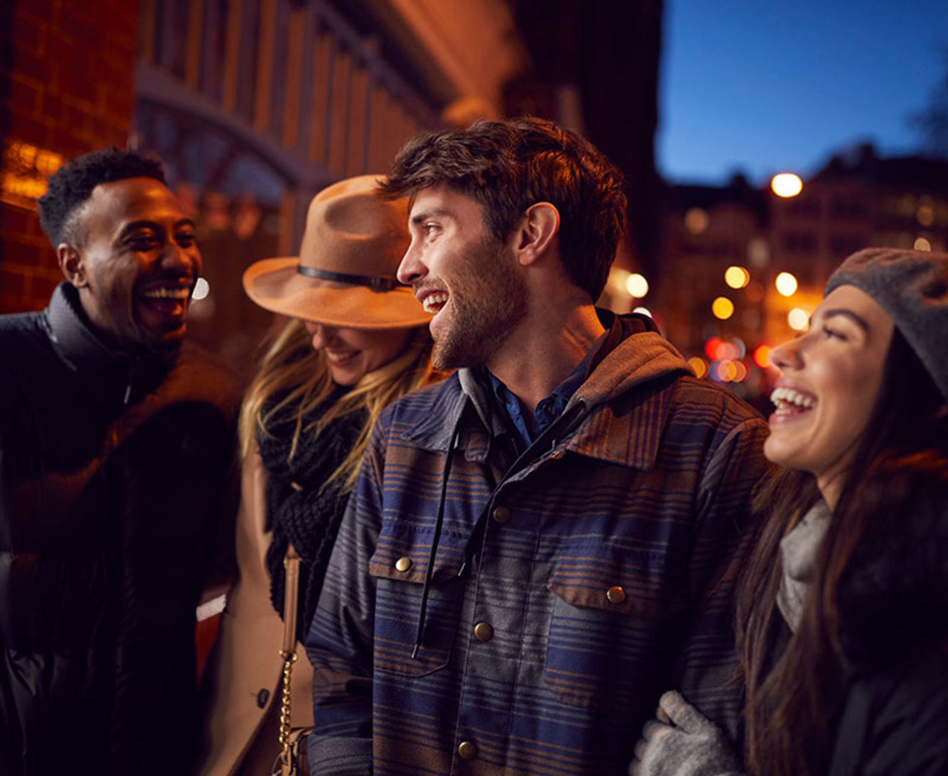Four friends laughing together on a chilly city sidewalk at night, bundled in coats, hats and scarves with warm bokeh streetlights in the background.