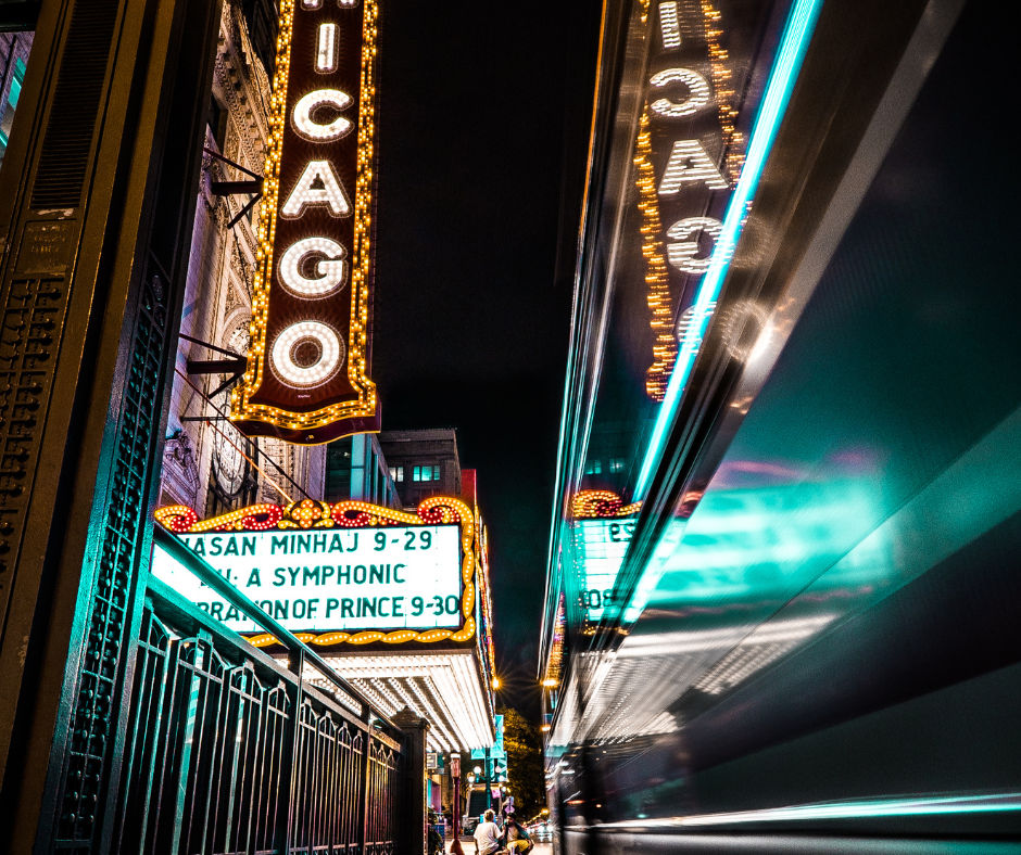 Nighttime Chicago theater marquee and vertical "CHICAGO" sign glowing above a downtown street with neon reflections and a motion-blurred passing bus.