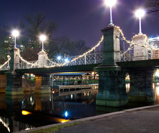 Ornate pedestrian suspension bridge draped in string lights over a calm river, illuminated lampposts and city skyline reflections at night in an urban park