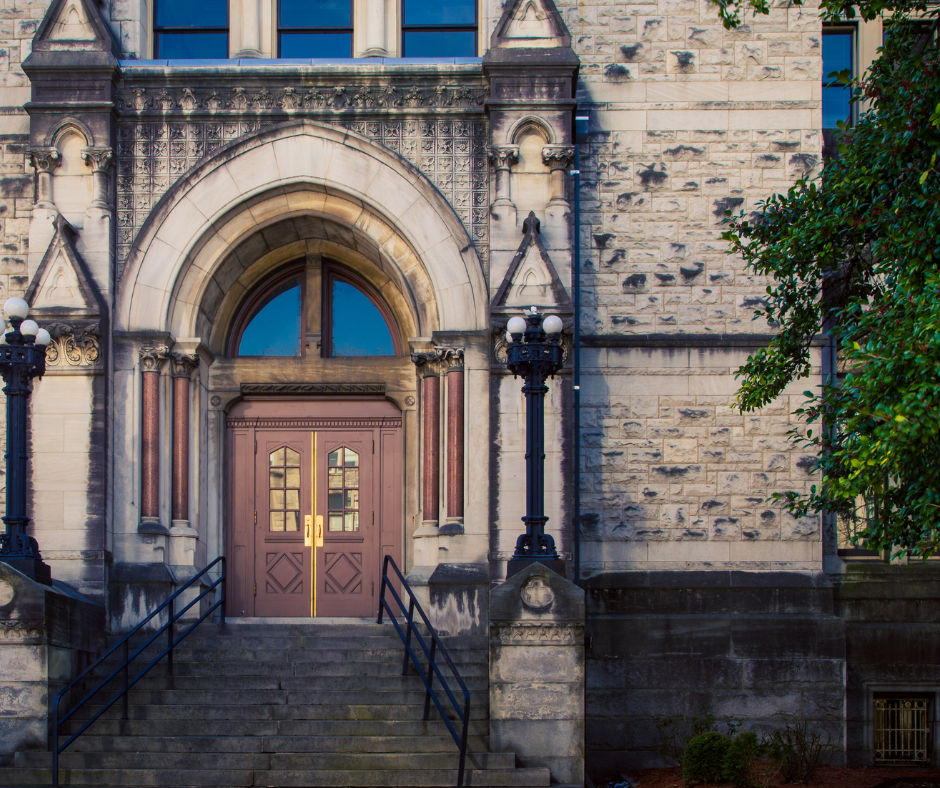 Historic stone building entrance with a grand arched doorway and double wooden doors with brass handles, wide stone steps flanked by ornate cast-iron lampposts and a leafy tree on the right.