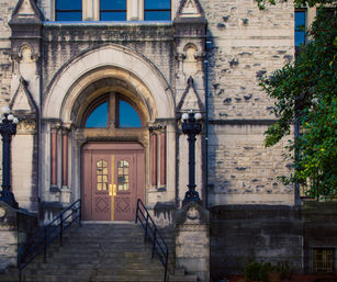 Historic stone building entrance with a grand arched doorway and double wooden doors with brass handles, wide stone steps flanked by ornate cast-iron lampposts and a leafy tree on the right.