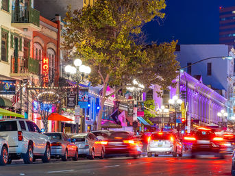 Vibrant downtown nightlife scene with glowing neon signs, historic storefronts, street lamps, outdoor dining umbrellas, parked cars and streaking taillight trails on a busy city street