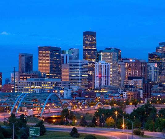 Twilight downtown skyline with glowing arched bridge, lit high-rise office towers, and tree-lined highway.