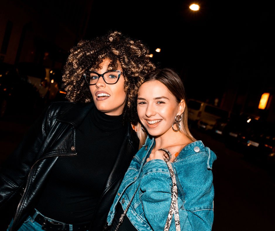 Two friends smiling on a city street at night — one with curly hair and glasses in a black leather jacket, the other in a denim jacket holding a patterned bag; candid urban nightlife portrait.