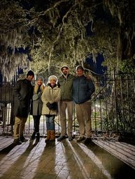 Five people in winter coats and hats posing at night in front of a wrought-iron gate beneath a large live oak draped with Spanish moss, long shadows cast on a wet brick walkway.