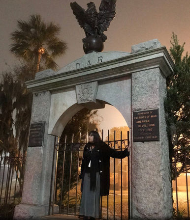 Person in a long coat standing at an eagle-topped granite arch and wrought-iron gate of a historic colonial cemetery at night, fog, palm trees, and a Revolutionary War memorial plaque.