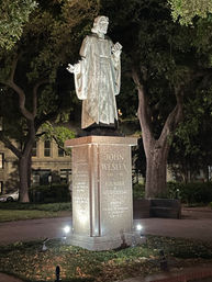 Nighttime view of an illuminated bronze statue of a robed clergyman on a granite pedestal with an engraved plaque, framed by large oak trees in a city park.