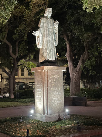 Nighttime view of an illuminated bronze statue of a robed clergyman on a granite pedestal with an engraved plaque, framed by large oak trees in a city park.