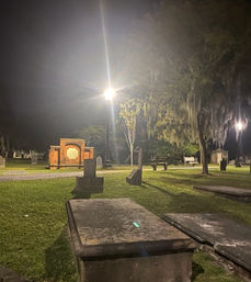 Nighttime view of a historic cemetery with stone ledger graves in the foreground, a glowing lamppost lighting a brick memorial, benches and Spanish moss-draped oak trees under an eerie, soft glow.