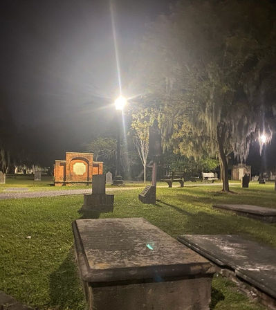 Nighttime view of a historic cemetery with stone ledger graves in the foreground, a glowing lamppost lighting a brick memorial, benches and Spanish moss-draped oak trees under an eerie, soft glow.