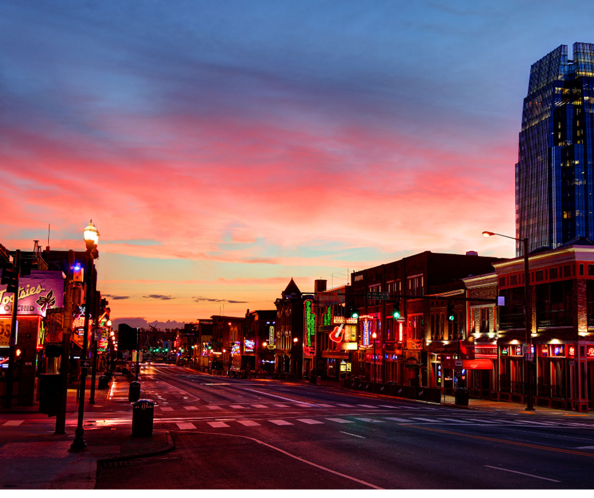 Vibrant downtown nightlife street at sunset — empty wet city road lined with neon-lit bars and restaurants, colorful pink-and-blue sky and a glass high-rise on the right.