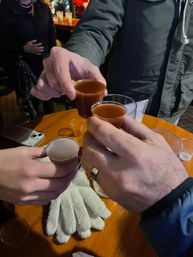 Four hands clinking small shot cups of brown liquor over a wooden bar table, knit gloves and a smartphone on the table, winter coats and a crowd blurred in the background