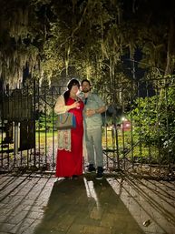 Couple at night — woman in a red dress and man in casual shirt holding drinks, posing before an ornate wrought-iron gate under Spanish-moss-draped live oaks at a historic park entrance.