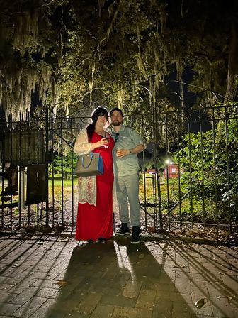 Couple at night — woman in a red dress and man in casual shirt holding drinks, posing before an ornate wrought-iron gate under Spanish-moss-draped live oaks at a historic park entrance.