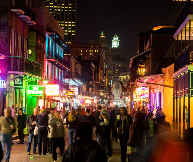 Nighttime New Orleans French Quarter scene on Bourbon Street — bustling crowd, colorful neon bar signs, wrought-iron balconies and historic buildings.