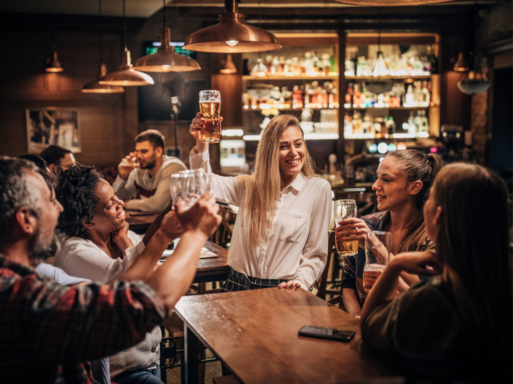Friends raising pints and cheering in a cozy wood-lit pub, smiling around a table under pendant lights with a stocked bar in the background.