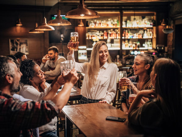 Friends raising pints and cheering in a cozy wood-lit pub, smiling around a table under pendant lights with a stocked bar in the background.