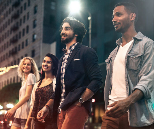 Four young adults walking together on a downtown city street at night, smiling under streetlights and tall buildings — upbeat urban nightlife scene.