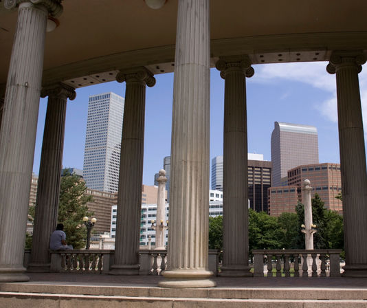 Classical stone colonnade framing the downtown Denver skyline with modern skyscrapers, leafy park trees, and a person sitting on a bench in the shade.