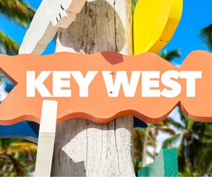 Bright orange wooden 'KEY WEST' sign on a weathered post with colorful sign pieces, palm trees and clear blue sky in the background for a sunny Florida Keys tropical vibe.