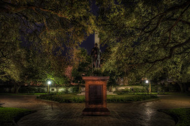 Nighttime historic park scene: a statue on a brick pedestal centered under a leafy tree canopy, glowing lampposts, cobblestone walkways radiating outward and a tall stone obelisk visible in the background.