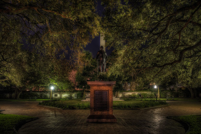 Nighttime historic park scene: a statue on a brick pedestal centered under a leafy tree canopy, glowing lampposts, cobblestone walkways radiating outward and a tall stone obelisk visible in the background.