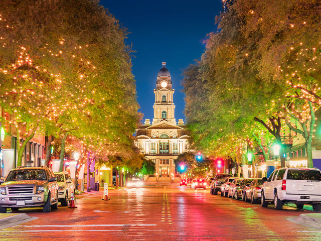 Downtown evening scene: tree-lined brick street draped in warm string lights leading to a historic courthouse clock tower, with parked cars and colorful streetlight reflections on the pavement.