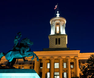 Dramatic night view of a historic neoclassical government building landmark with a glowing cupola and columned façade, foreground bronze equestrian statue silhouetted against a deep blue sky.