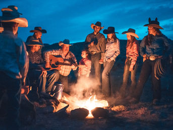 Cowboy sing-along at twilight: group of cowboys and cowgirls in hats and boots gathered around a ranch campfire, a man strumming an acoustic guitar while others listen, with a coffee pot and lariat nearby.