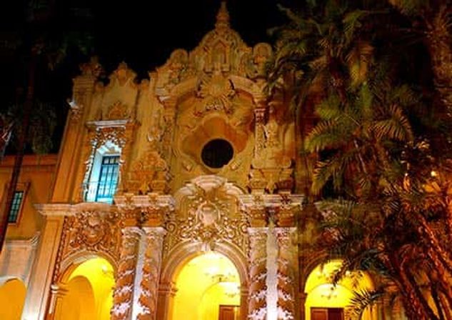 Historic Spanish Baroque-style facade aglow at night, ornate carved columns and arches framed by palm trees and warm yellow lighting.
