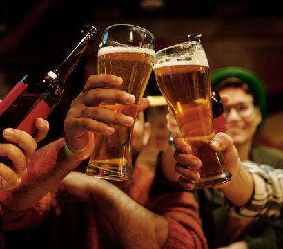 Friends clinking tall amber beer glasses and a bottle in a cozy neighborhood pub, foamy craft beer raised in a celebratory toast.