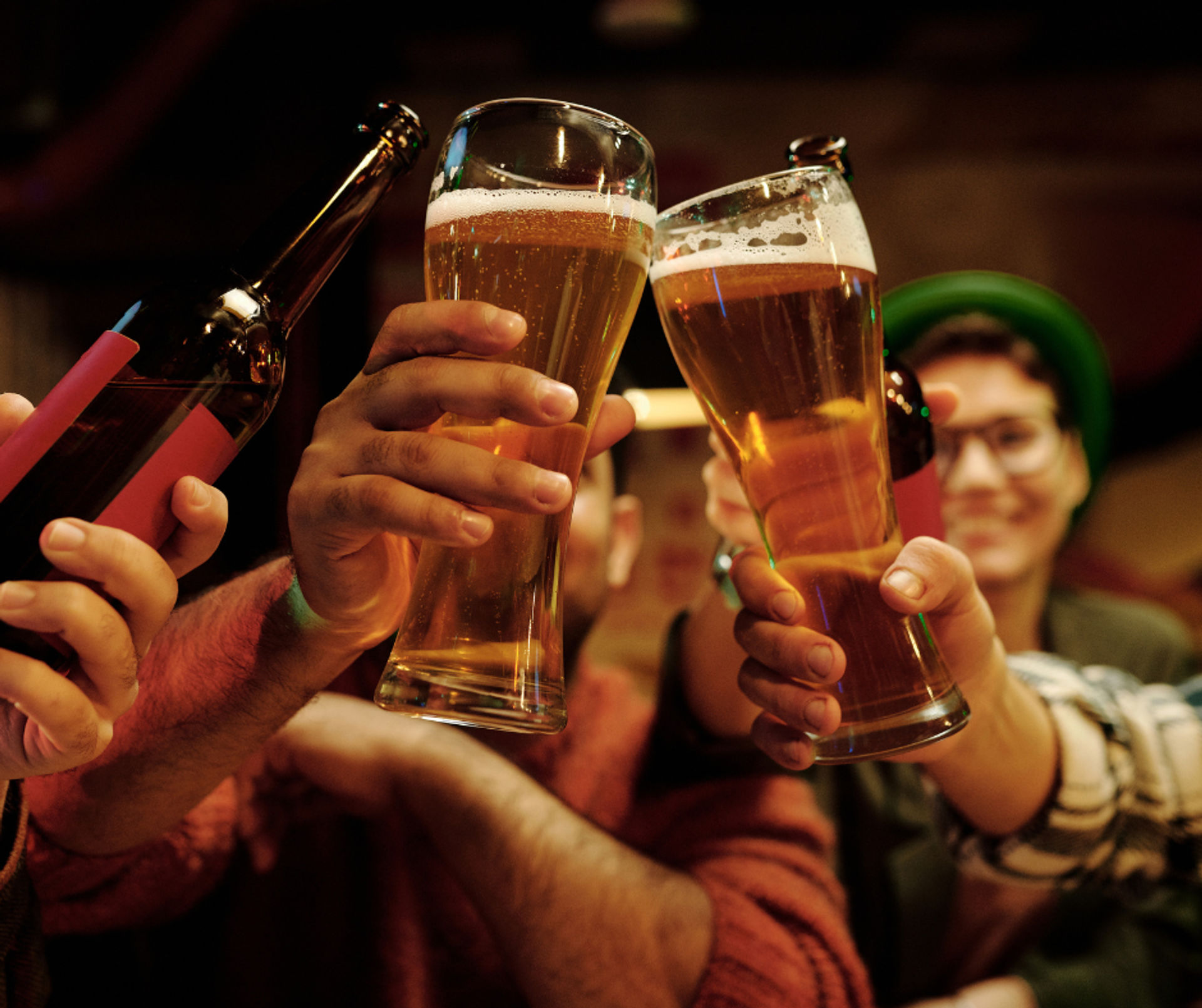 Friends clinking tall amber beer glasses and a bottle in a cozy neighborhood pub, foamy craft beer raised in a celebratory toast.