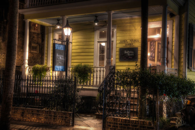Nighttime scene of a yellow historic porch lit by a warm streetlamp, with white columns, hanging ferns, ornate wrought-iron gate and brick steps.