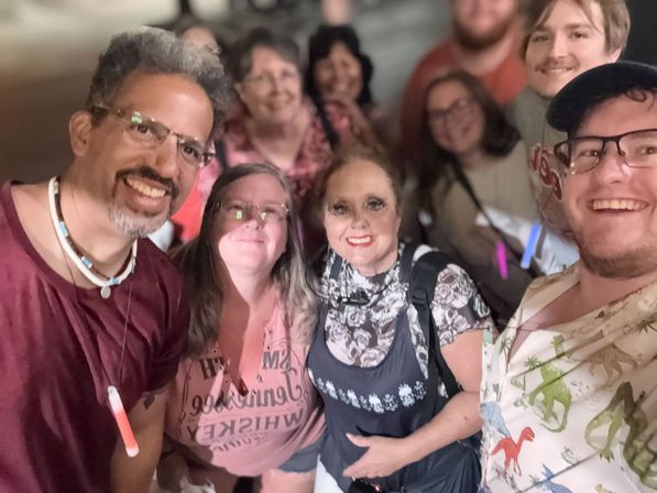 Smiling group selfie of friends at a lively nighttime street festival, close-up of diverse adults with glow sticks and blurred city lights in the background.