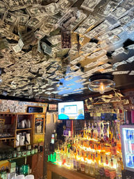 Cozy bar interior with ceiling covered in signed one-dollar bills, glowing backlit liquor bottles, a glass pendant light and a TV near an exit sign.