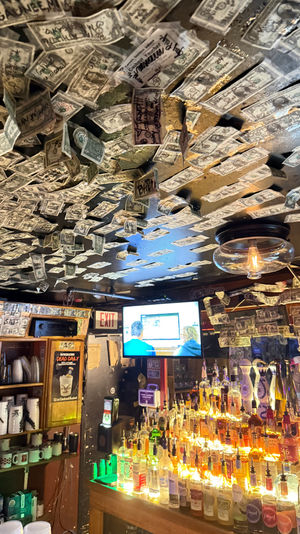 Cozy bar interior with ceiling covered in signed one-dollar bills, glowing backlit liquor bottles, a glass pendant light and a TV near an exit sign.