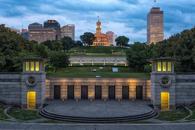 Dusk city skyline with an illuminated historic state capitol on a tree-lined hill, modern office towers behind, and a lit stone terraced amphitheater and plaza in the foreground.